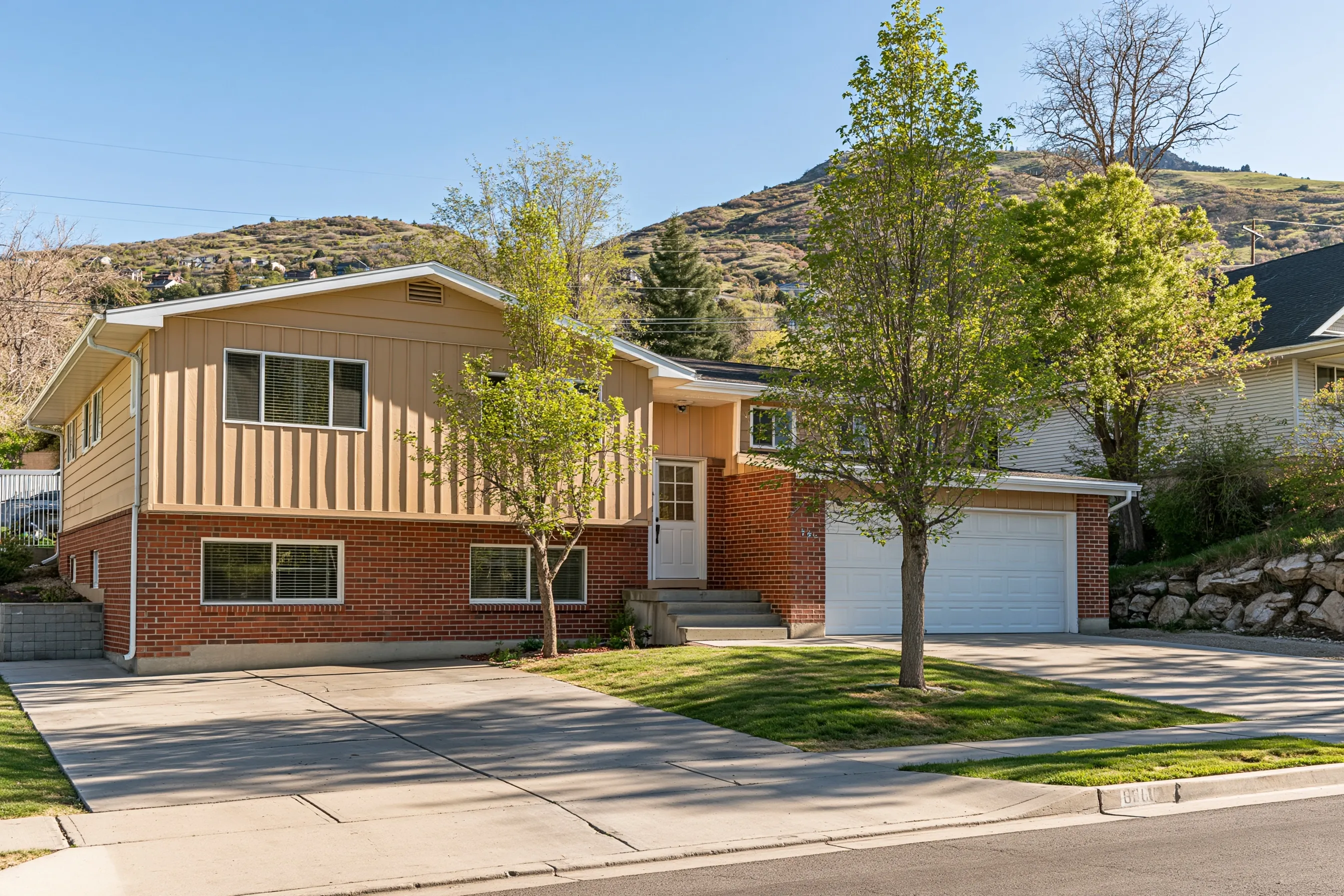 Split-entry house from the 1970s with a brick lower level, light siding above, basement windows half above grade, a two-car garage and the Wasatch foothills behind