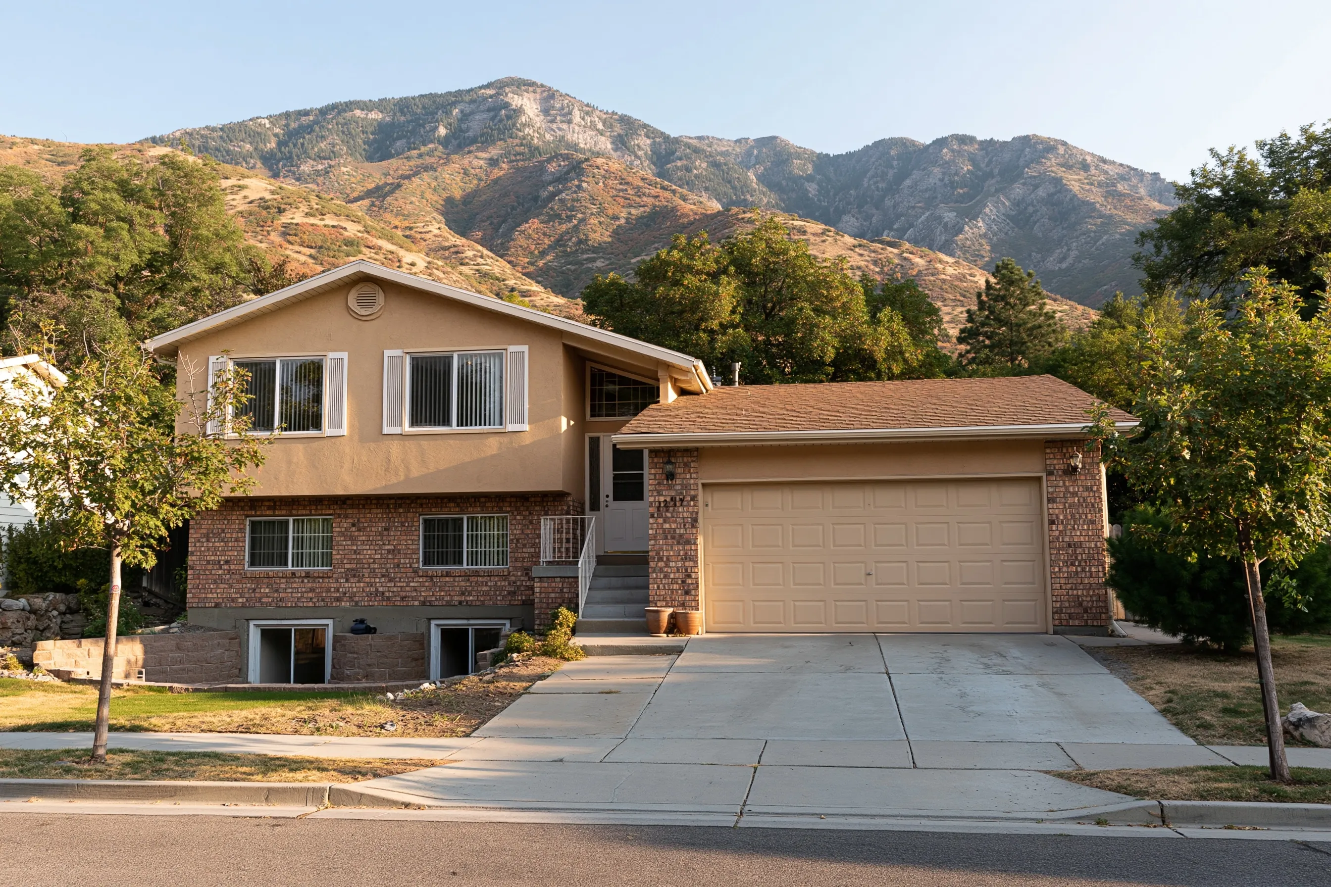 Split-level house with a tan stucco upper floor, a brick lower floor, two window wells at the foundation, a two-car garage and a mountain behind